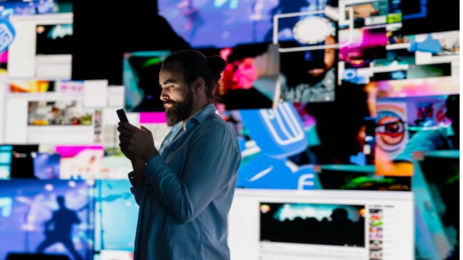 A imagem mostra um homem de perfil, com cabelo apanhado num coque e barba, a olhar atentamente para o ecrã do seu smartphone. Traja uma camisa azul clara e está posicionado num ambiente escuro, cercado por múltiplos ecrãs e painéis digitais iluminados.