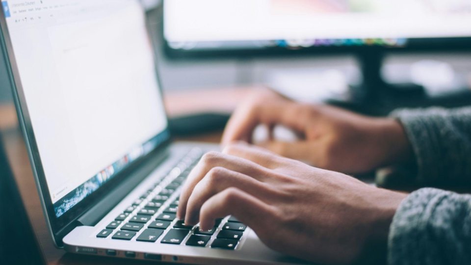 As mãos estão posicionadas sobre o teclado preto com estrutura de alumínio prata de um laptop (estilo MacBook), simulando a digitação de um texto.